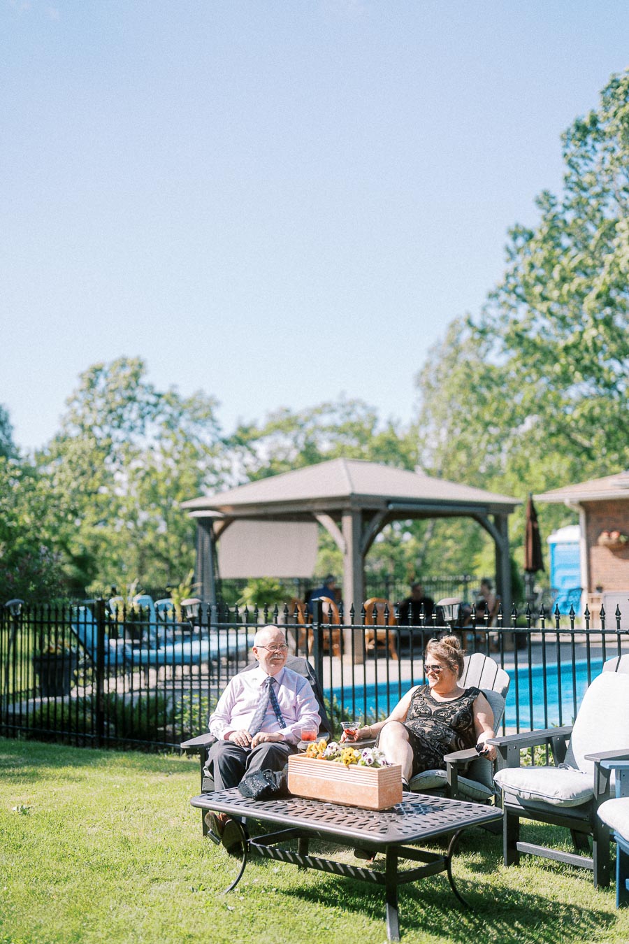 Elderly couple relaxing on patio chairs by a poolside in a sunny backyard setting, with a flower box centerpiece and a gazebo in the background.