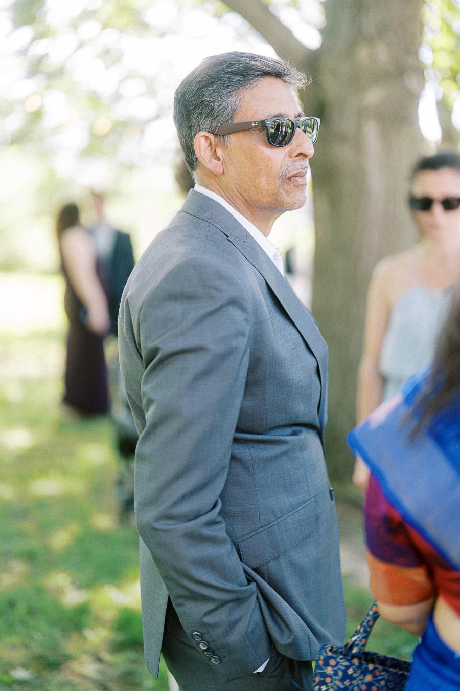 Man in a gray suit and sunglasses standing outdoors at a social gathering, with sunlight filtering through trees in the background.