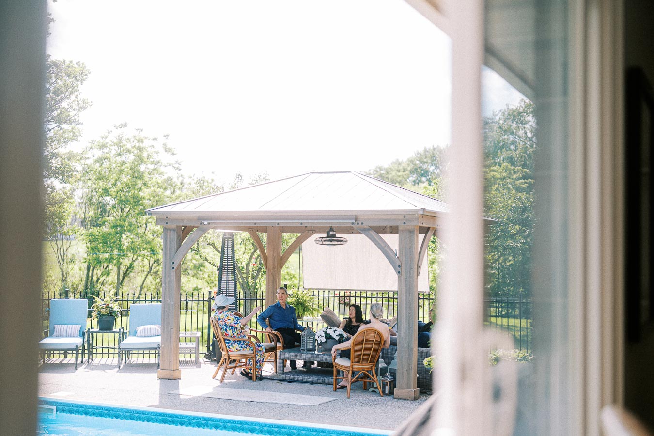 Group of people relaxing under a wooden gazebo by a swimming pool, enjoying a sunny day with greenery in the background.