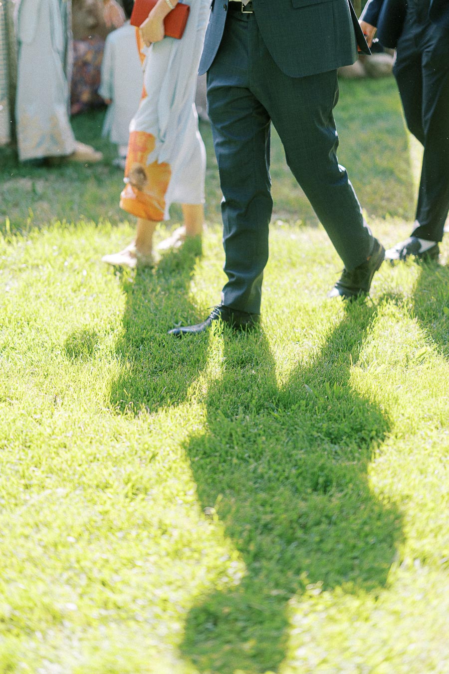 A group of people walking on sunny green grass, casting long shadows, with one person in a blue suit leading the way, suggesting a formal outdoor event.