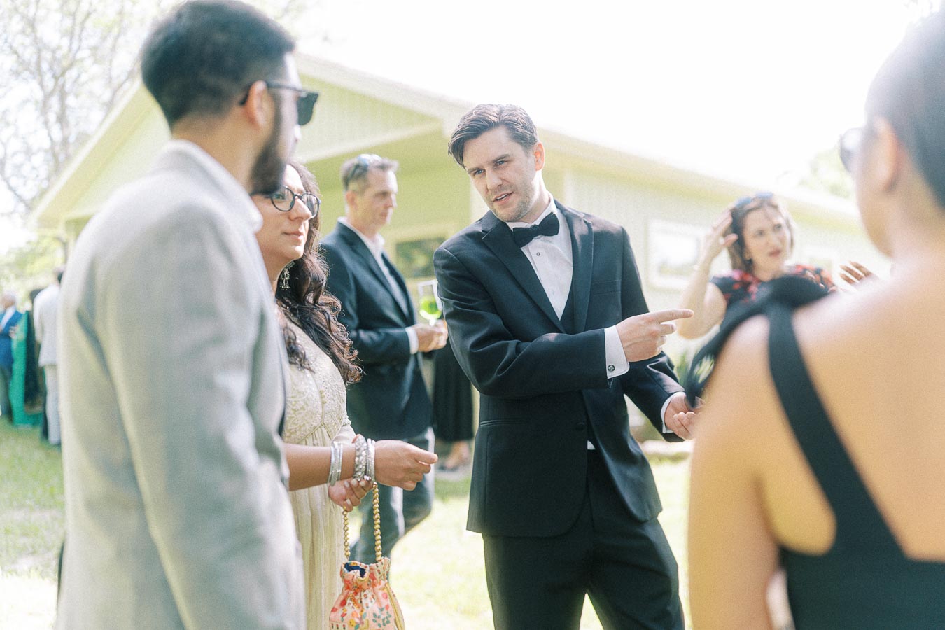 Group of people in formal attire socializing outdoors at an event; a man in a tuxedo is engaging in conversation, setting a sophisticated and stylish atmosphere.