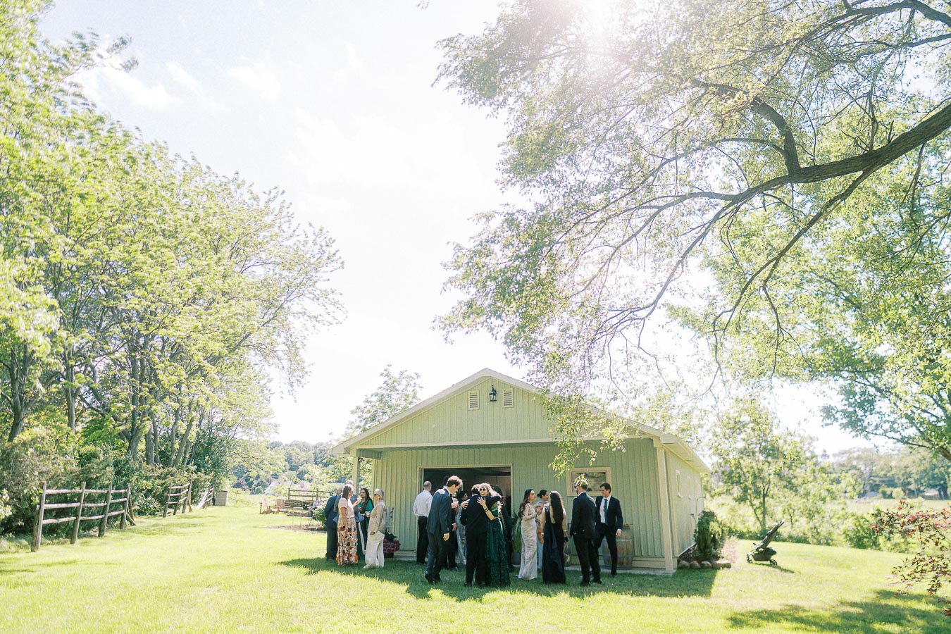 Group of people socializing outdoors at a wedding reception near a rustic barn in a lush green landscape, with trees and a bright sky.