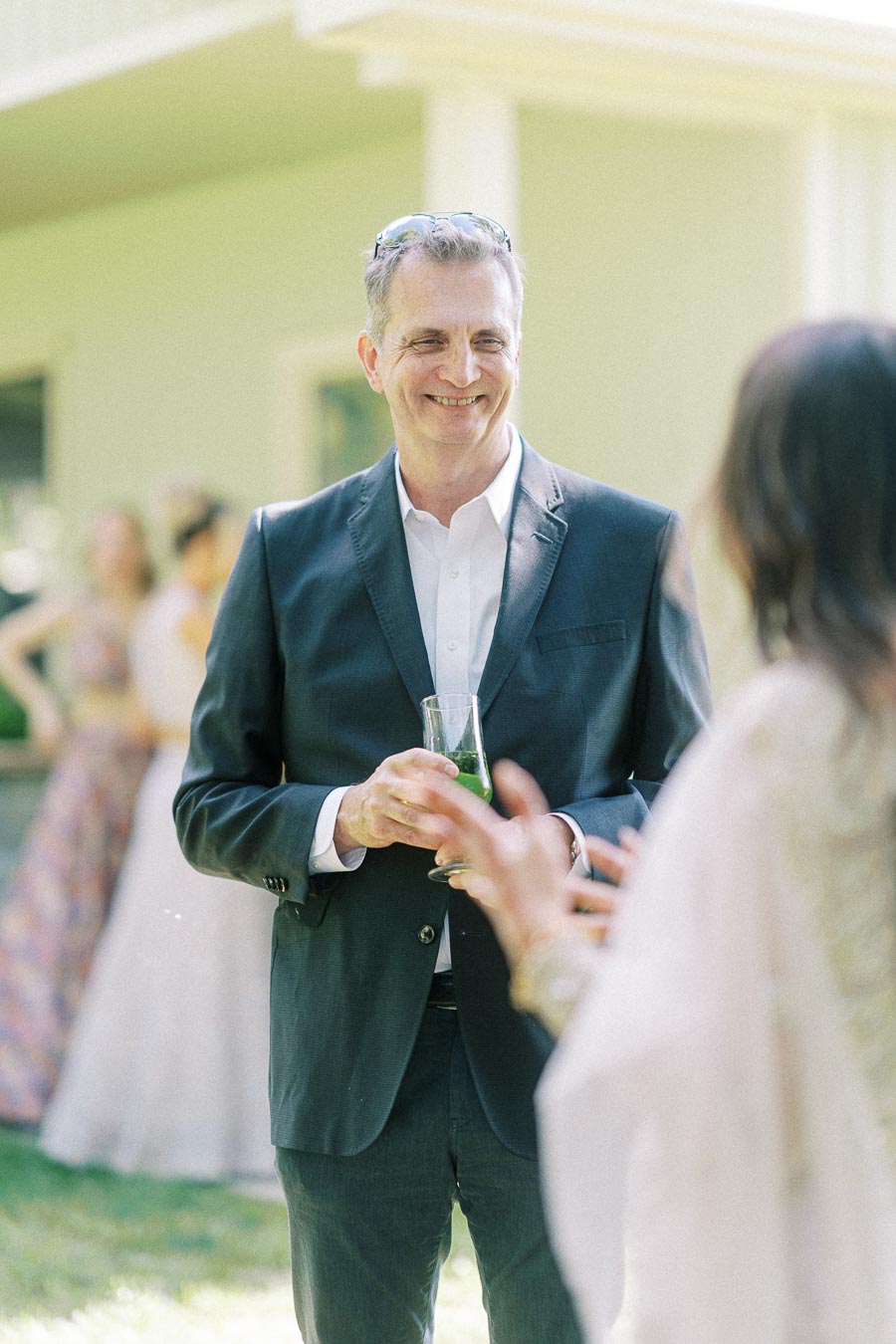 Man in a dark suit smiling and holding a drink while conversing with a woman outdoors at a daytime gathering, with a blurred background.