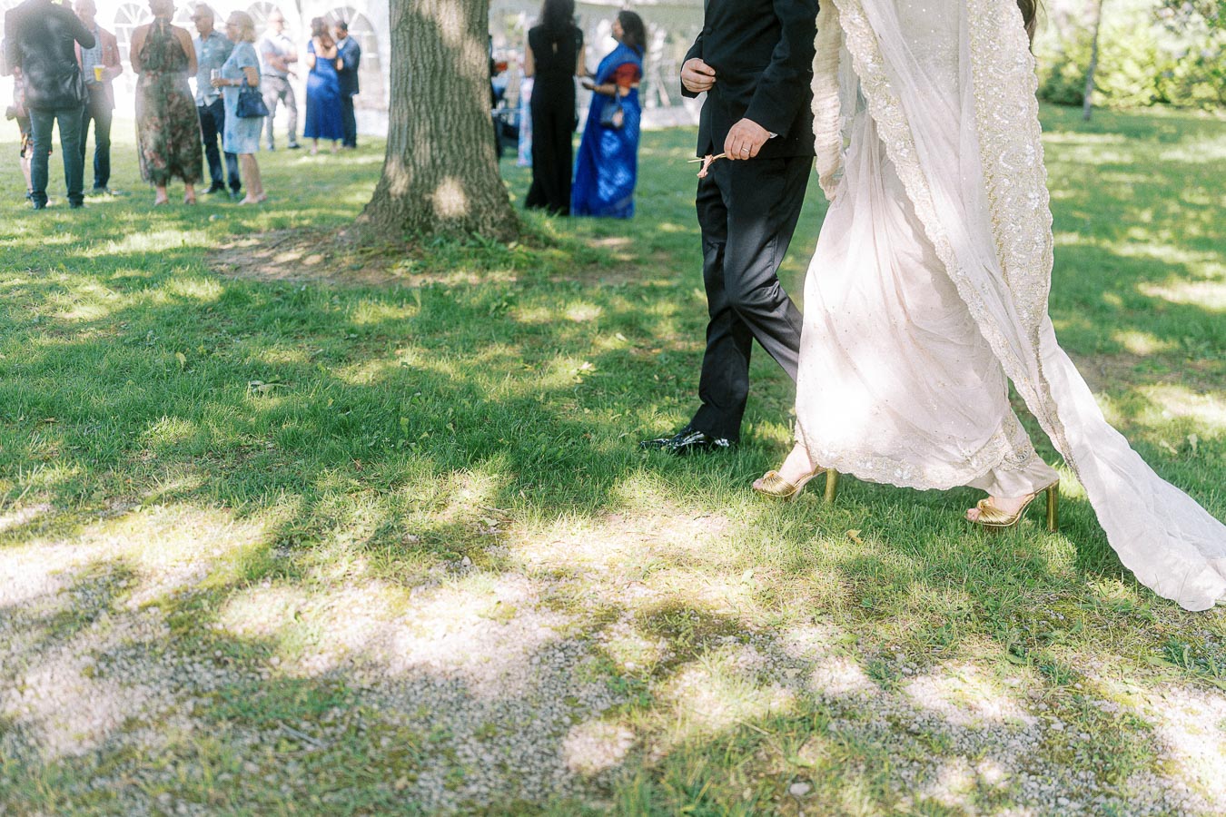 Elegant couple walking on grass at an outdoor event; woman in an embroidered dress and gold heels, man in a black suit, with guests and trees in the background.