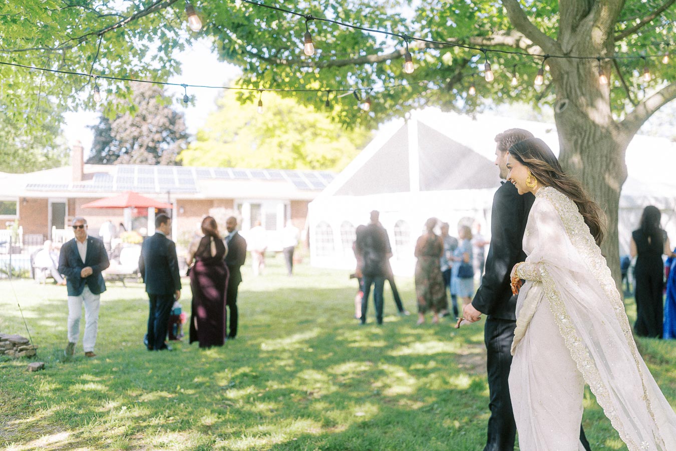 Elegant outdoor wedding reception with guests mingling under string lights, featuring a well-dressed couple walking hand in hand, surrounded by greenery and a white event tent in the background.