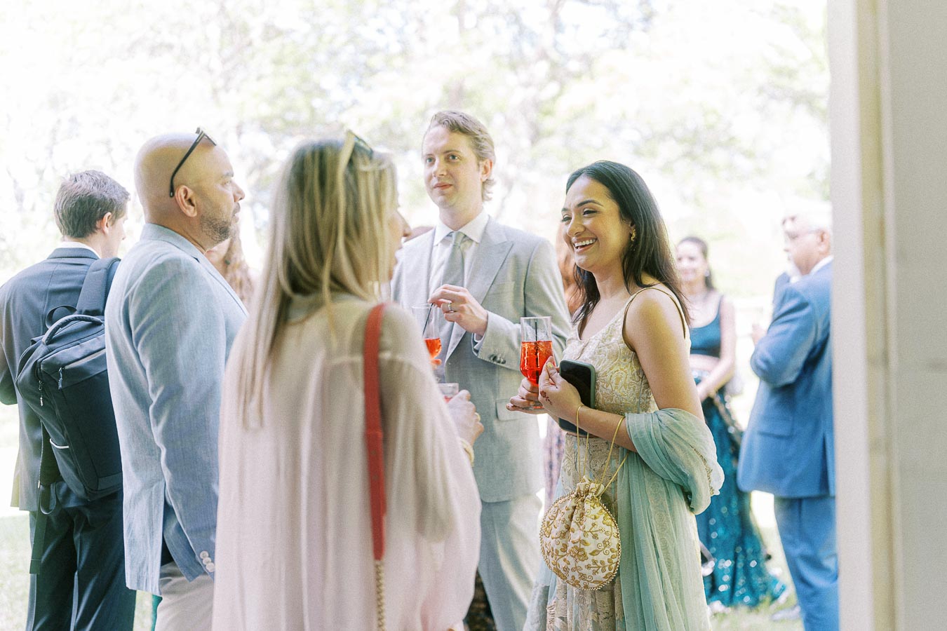 A group of elegantly dressed guests socializing outdoors at a wedding reception, holding glasses of red cocktails and dressed in formal attire, with a blurred natural background.