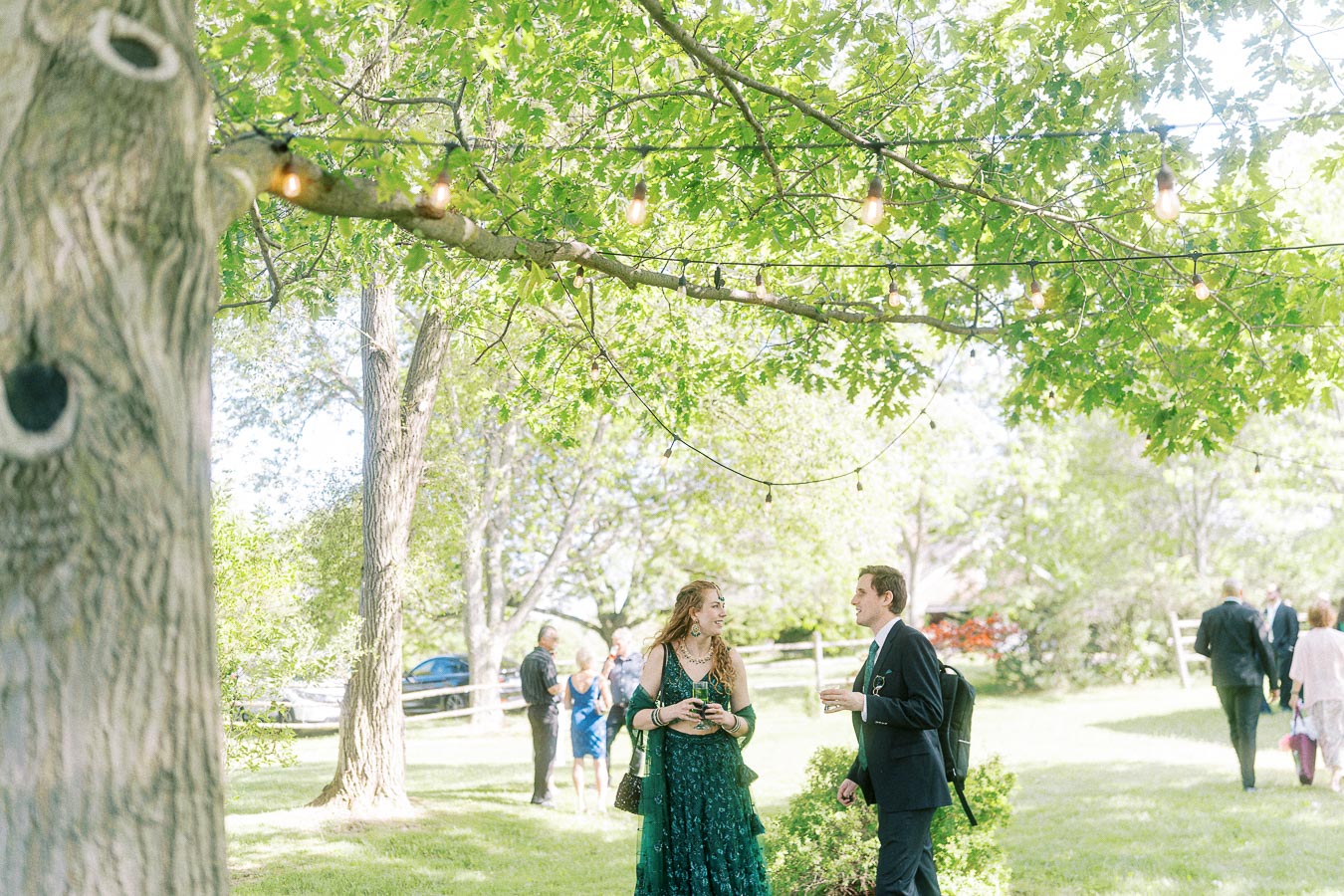 A man and woman converse at an outdoor event under string lights and lush green trees, with other guests mingling in the sunlit background.
