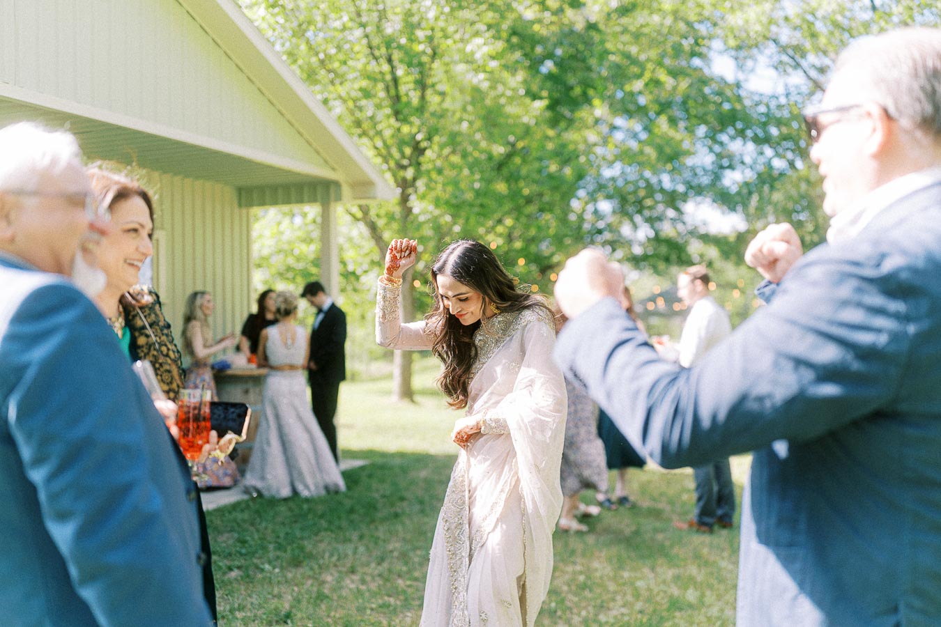 A joyful group of people enjoying an outdoor wedding celebration, with one woman in a traditional outfit dancing against a backdrop of trees and a sunny day.