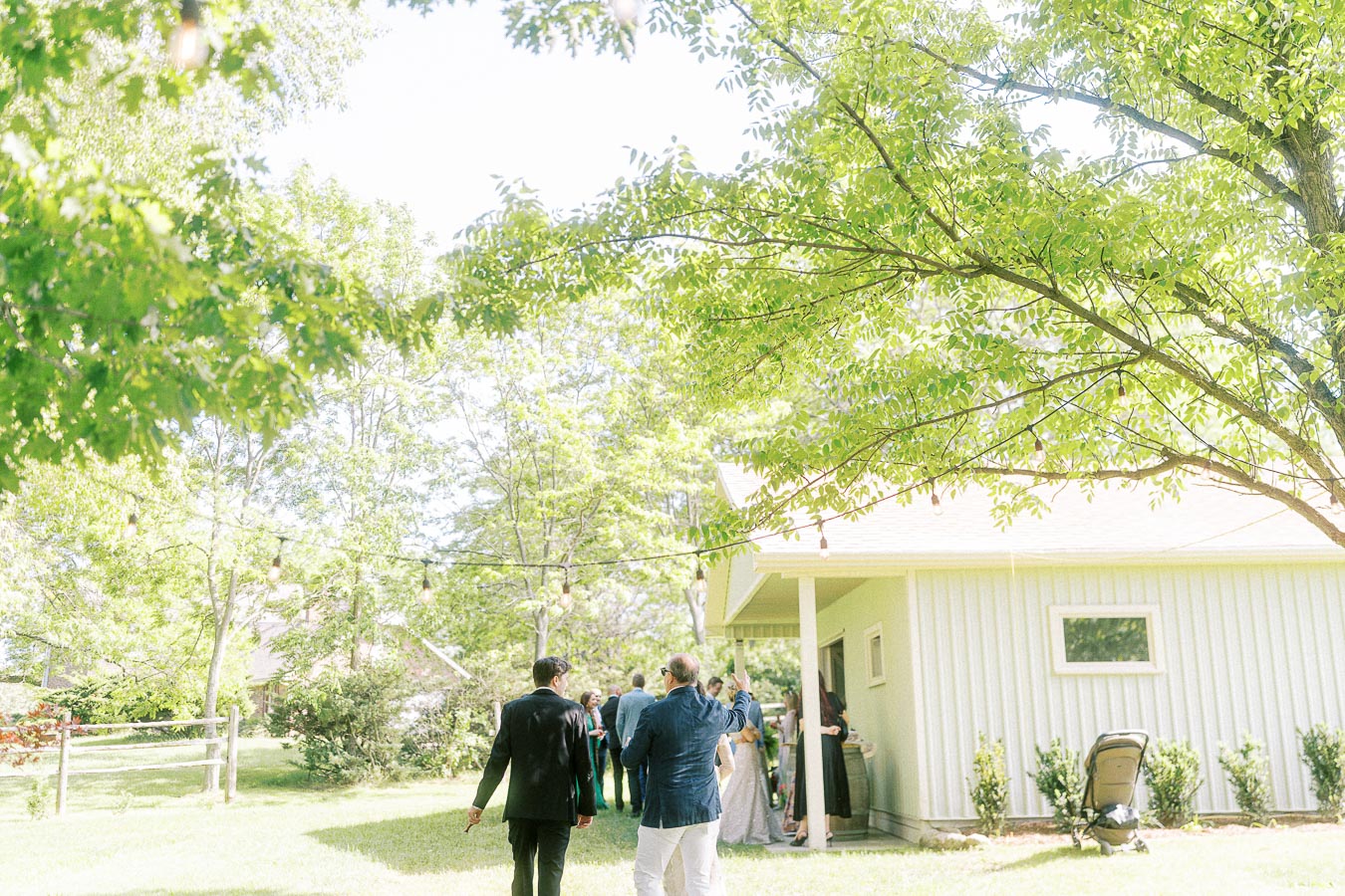 Group of people gathering outside a house surrounded by lush greenery and trees under bright sunlight.