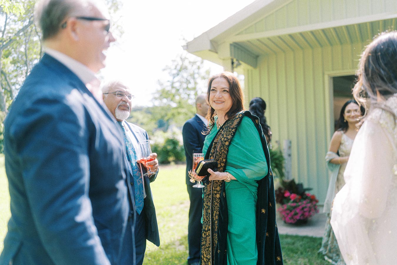 Group of people in formal attire enjoying an outdoor gathering, with a woman in a green saree smiling and holding a drink. The setting is a sunny garden with a building in the background.