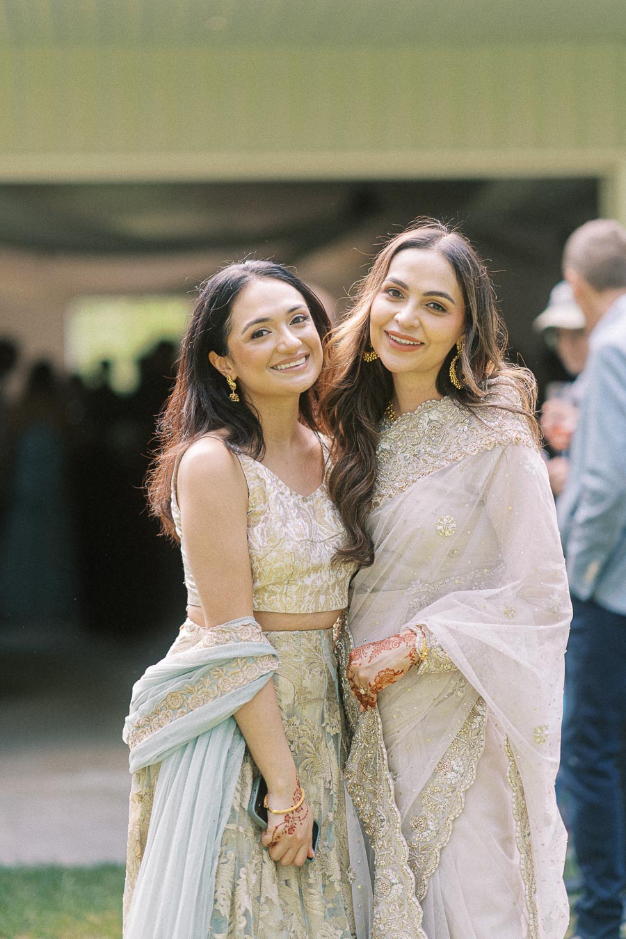 Two women dressed in elegant traditional attire, smiling outdoors at a festive event.