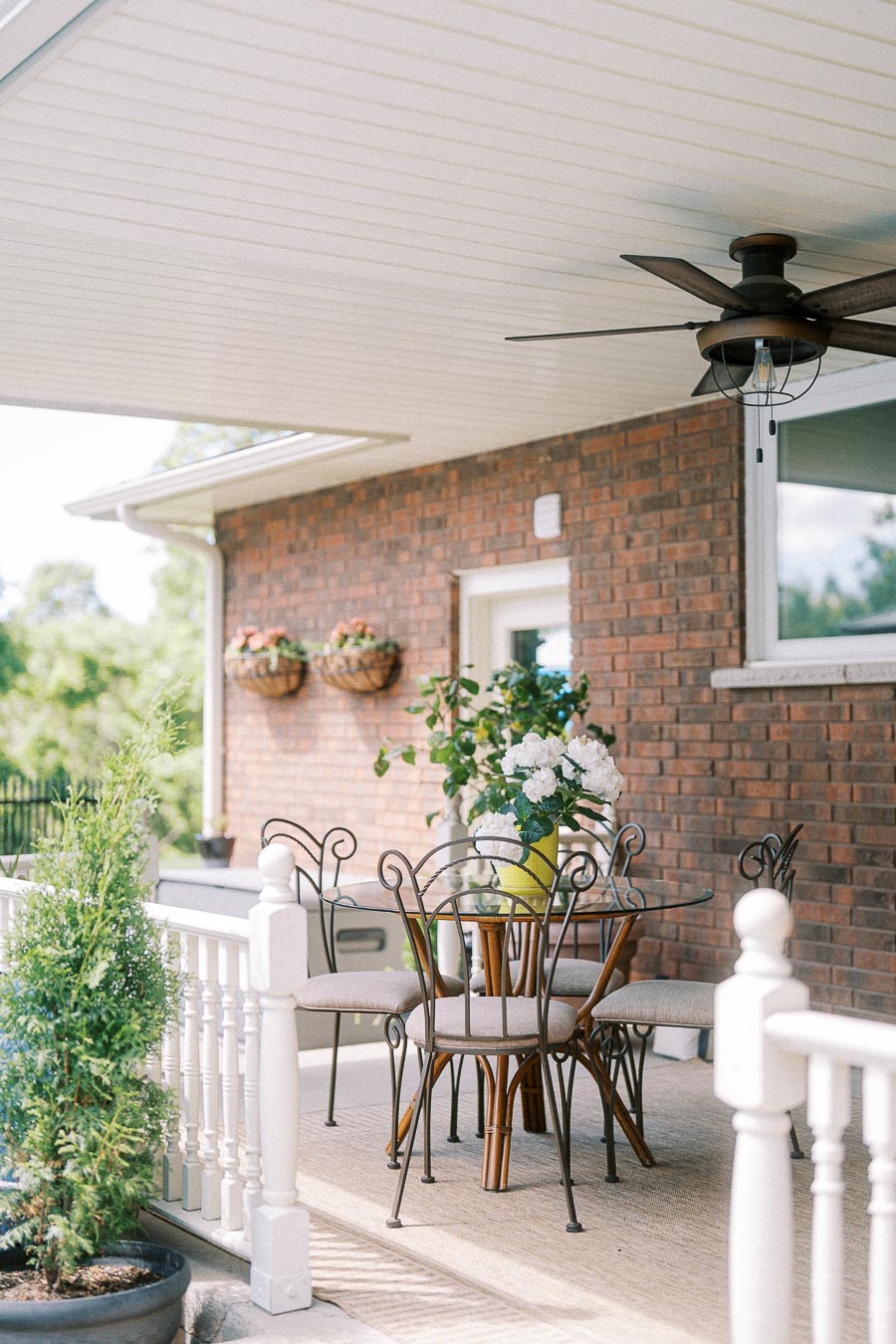Outdoor patio scene with a glass table and metal chairs adorned with a floral centerpiece. The brick wall background features two hanging planters, and a ceiling fan is mounted above, creating a cozy atmosphere.