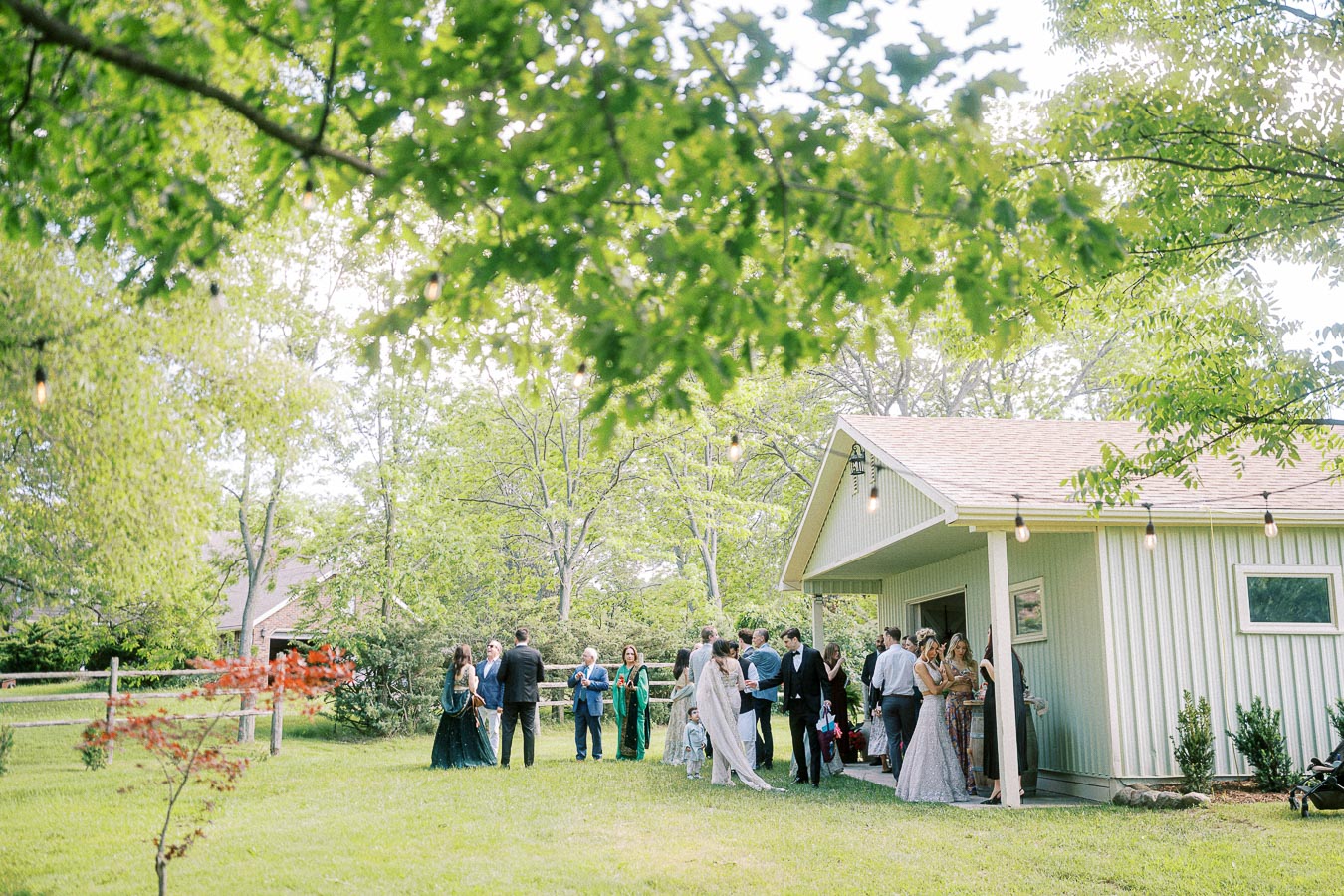 Outdoor wedding reception at a rustic venue with guests mingling beside a charming barn, surrounded by lush green trees and string lights, creating a serene and festive atmosphere.