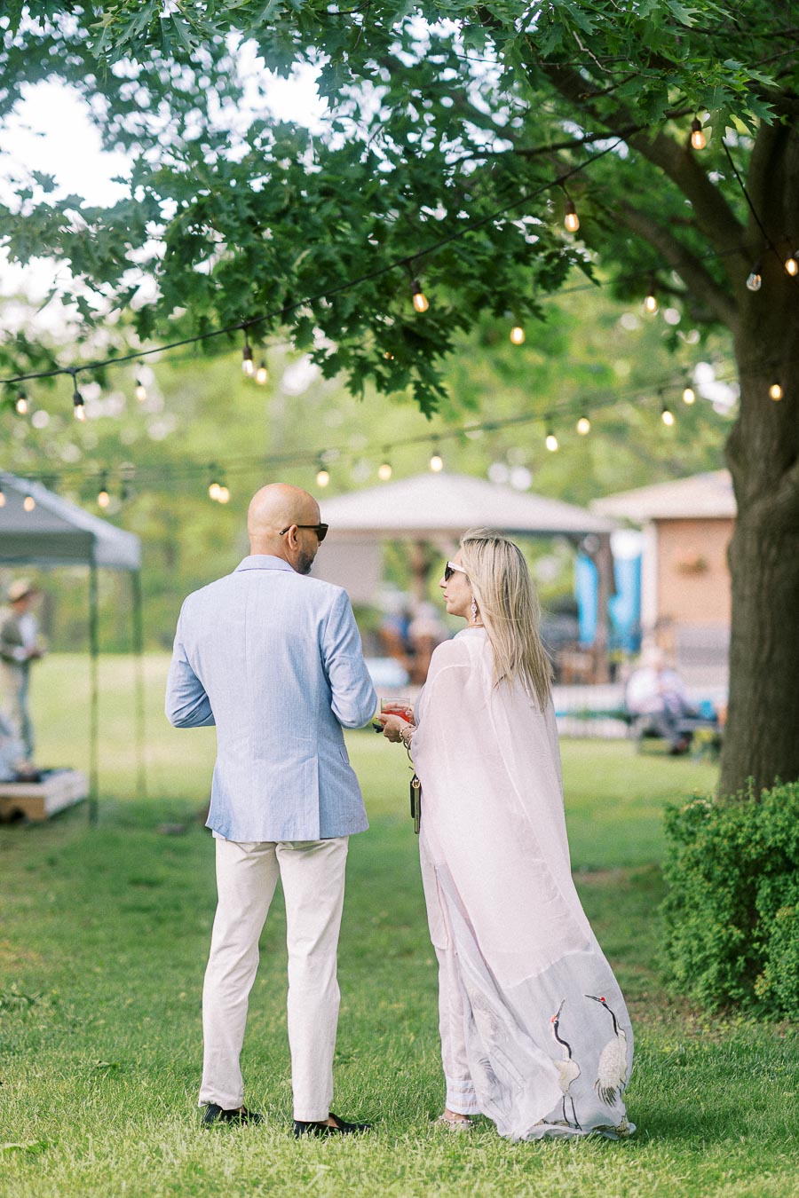 A stylish couple, dressed in light summer attire, enjoy a leisurely outdoor gathering under string lights and lush green trees. The scene captures a relaxed, elegant garden party atmosphere.