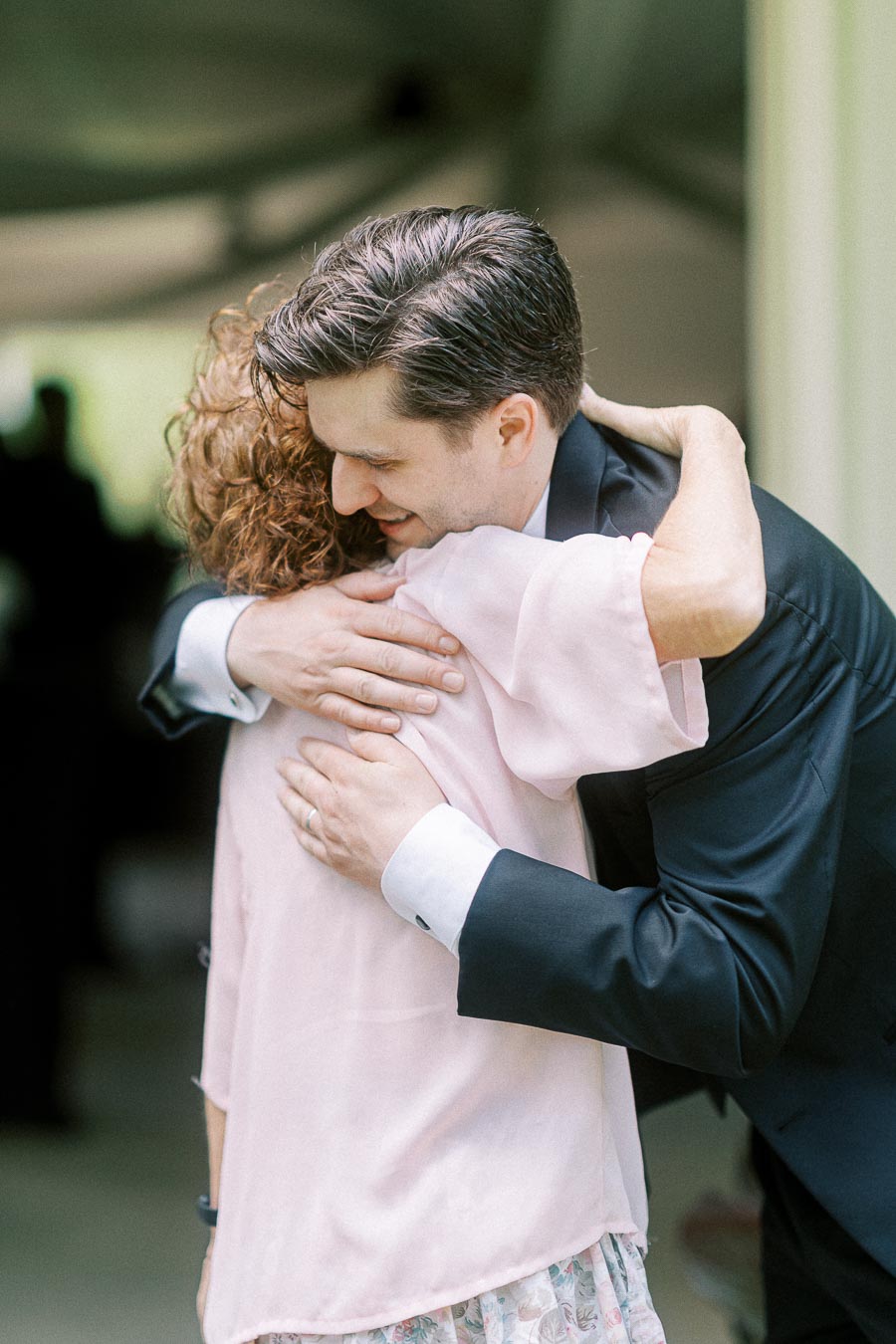 A young man in a black suit embraces a woman in a pink blouse during a heartfelt moment at a formal event.