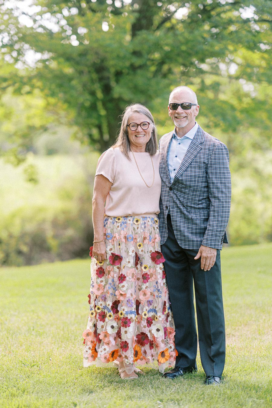 A smiling couple standing outdoors on a sunny day, with the woman wearing a floral skirt and the man dressed in a checkered blazer, set against a lush green background.