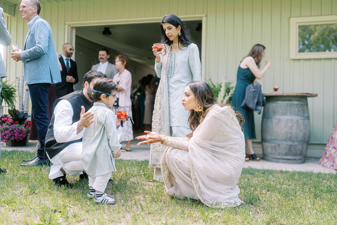 A joyful family gathering outdoors with people dressed in traditional South Asian attire; a woman kneels to interact with a small child, while others mingle in the background, enhancing the sense of community and celebration.