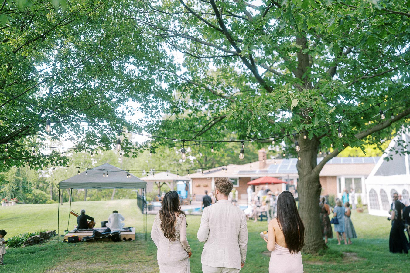 A serene outdoor garden party with guests dressed in light attire, approaching a canopy tent where musicians perform. Tree branches with string lights create a cozy atmosphere, perfect for a summer gathering.