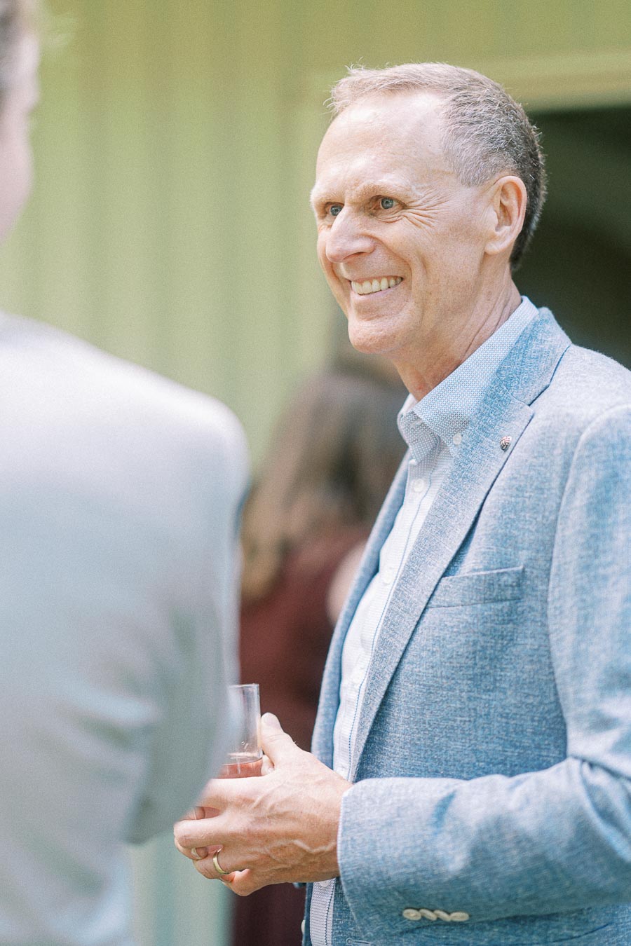 Elderly man in a blue blazer smiling and holding a drink during a social event, with blurred people in the background.