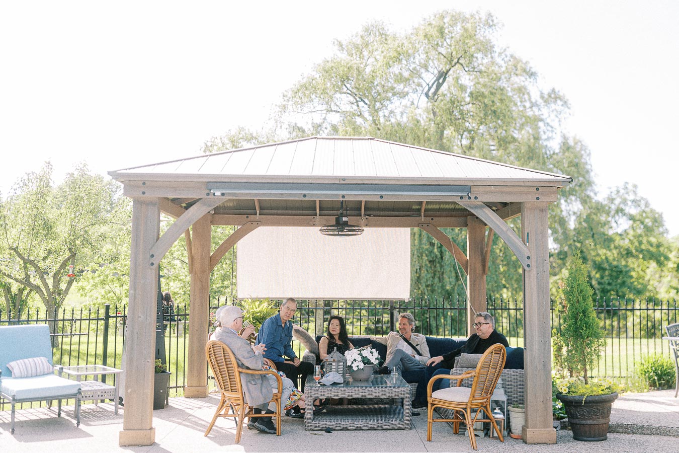 Group of people relaxing under a gazebo in a scenic outdoor setting, surrounded by lush greenery and garden furniture.
