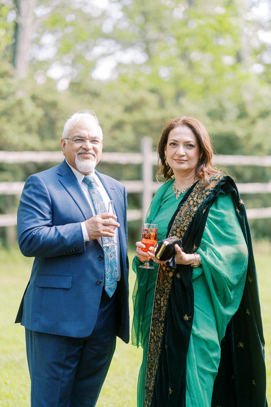 Elderly couple dressed in formal attire, standing outdoors with drinks in hand, surrounded by vibrant greenery.