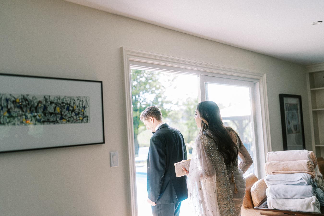 Elegant couple dressed for a formal occasion, with the woman in a beautifully embellished sari and the man in a suit, walking towards an open glass door leading to a bright outdoor area, with decorative artwork on the wall and neatly folded blankets on a nearby table.