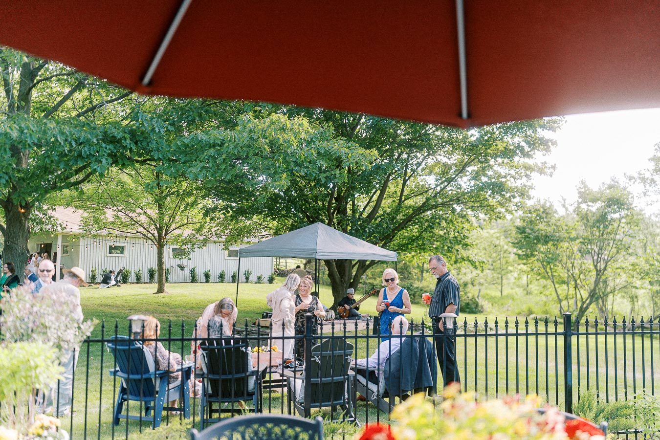 Outdoor garden party with people socializing under a canopy in a lush green backyard setting, featuring a wooden house, a large umbrella, and vibrant foliage in the foreground.