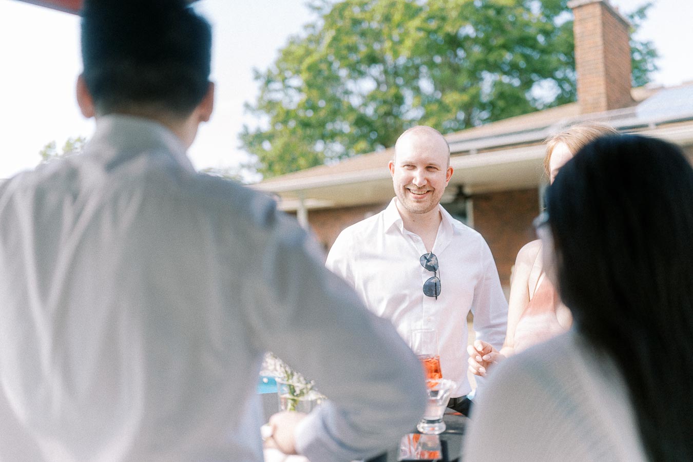 Group of people enjoying an outdoor gathering, smiling and conversing around a table with drinks, wearing casual summer attire in a sunny setting.
