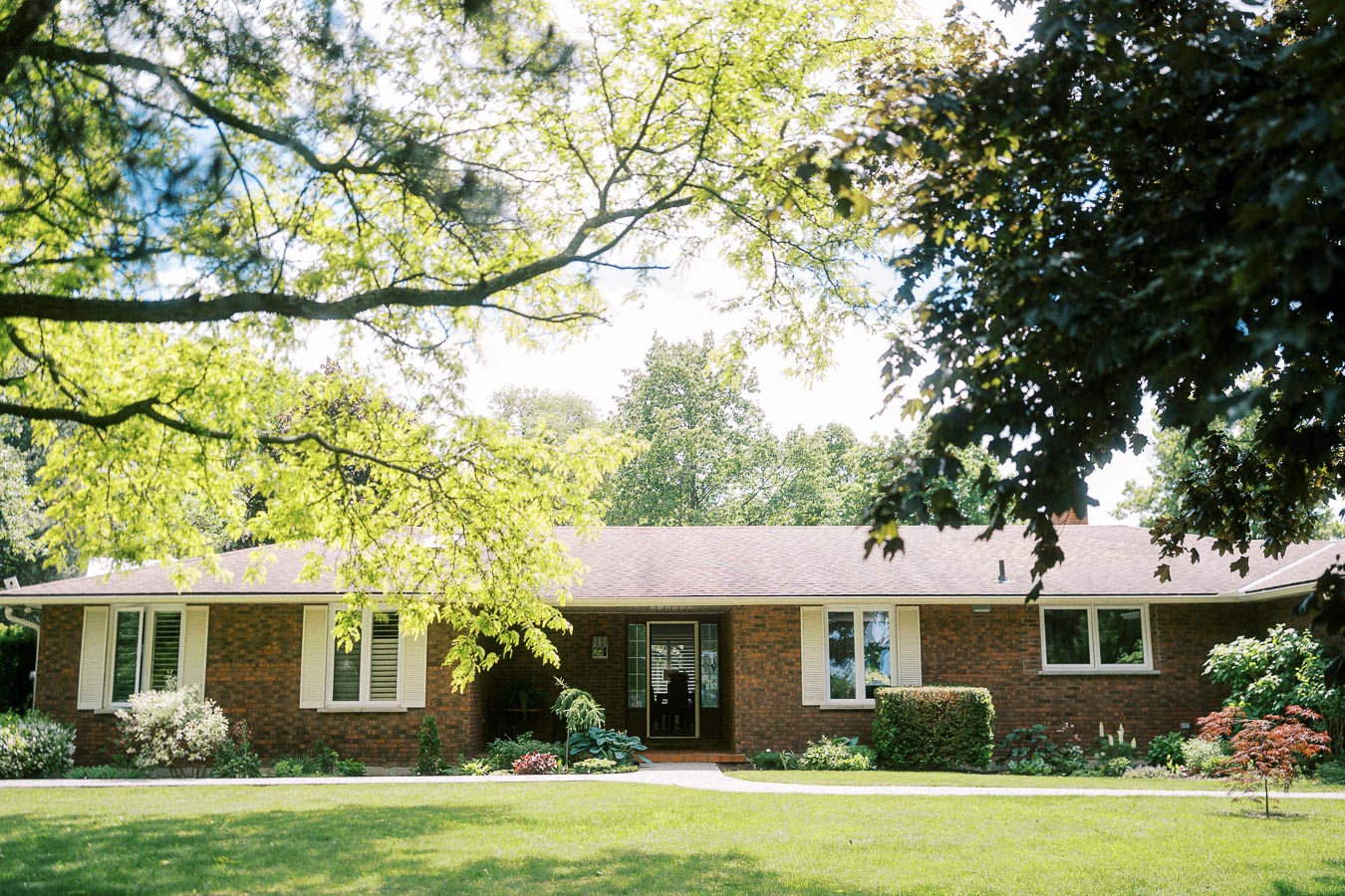Single-story brick house surrounded by lush green lawn and trees, under a sunny, clear sky.