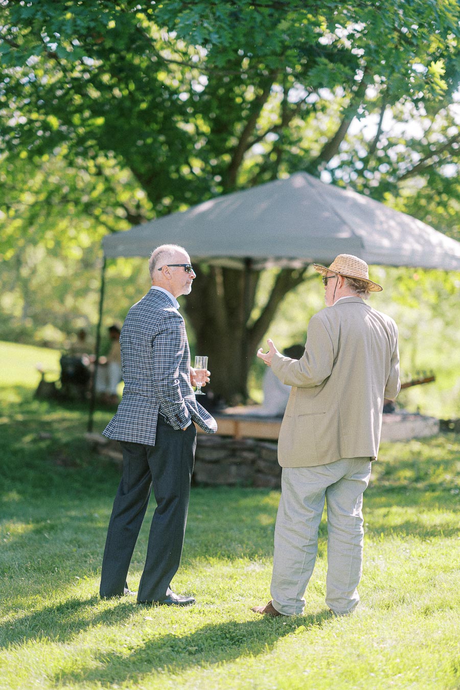 Two men in conversation outdoors at a garden party, dressed in suits and hats, standing on a sunny lawn with a tree and canopy in the background.