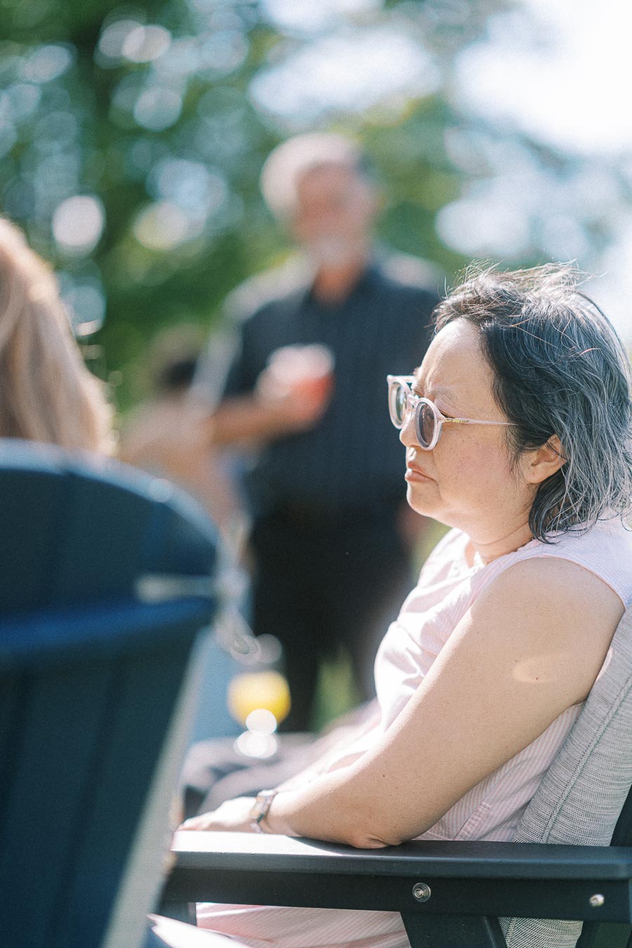 A woman wearing sunglasses sitting on a patio chair outdoors, with blurred people and lush greenery in the background, enjoying a sunny day.