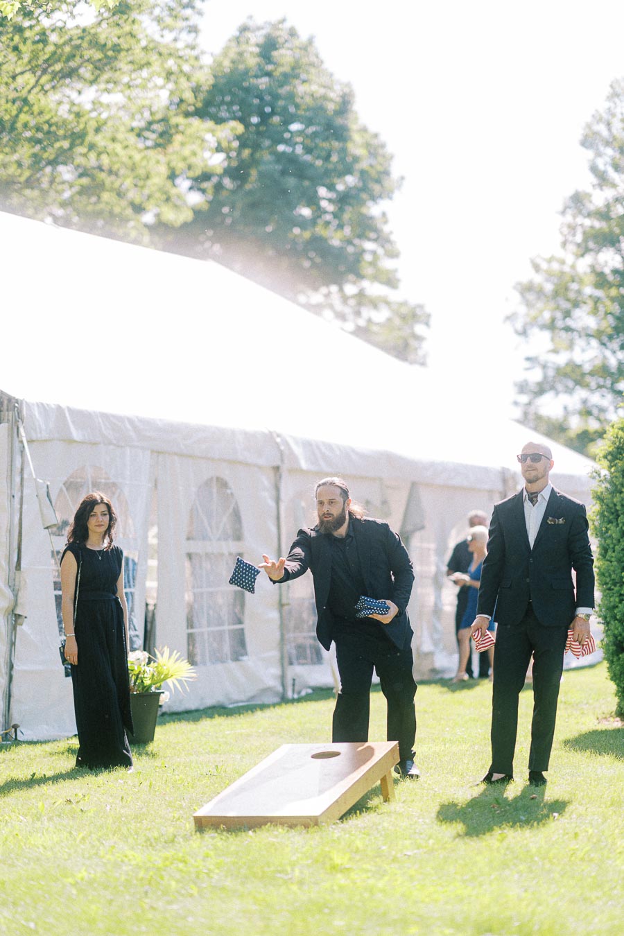 Men playing cornhole at an outdoor event near a white tent, dressed in formal attire, under a sunny, tree-lined setting.