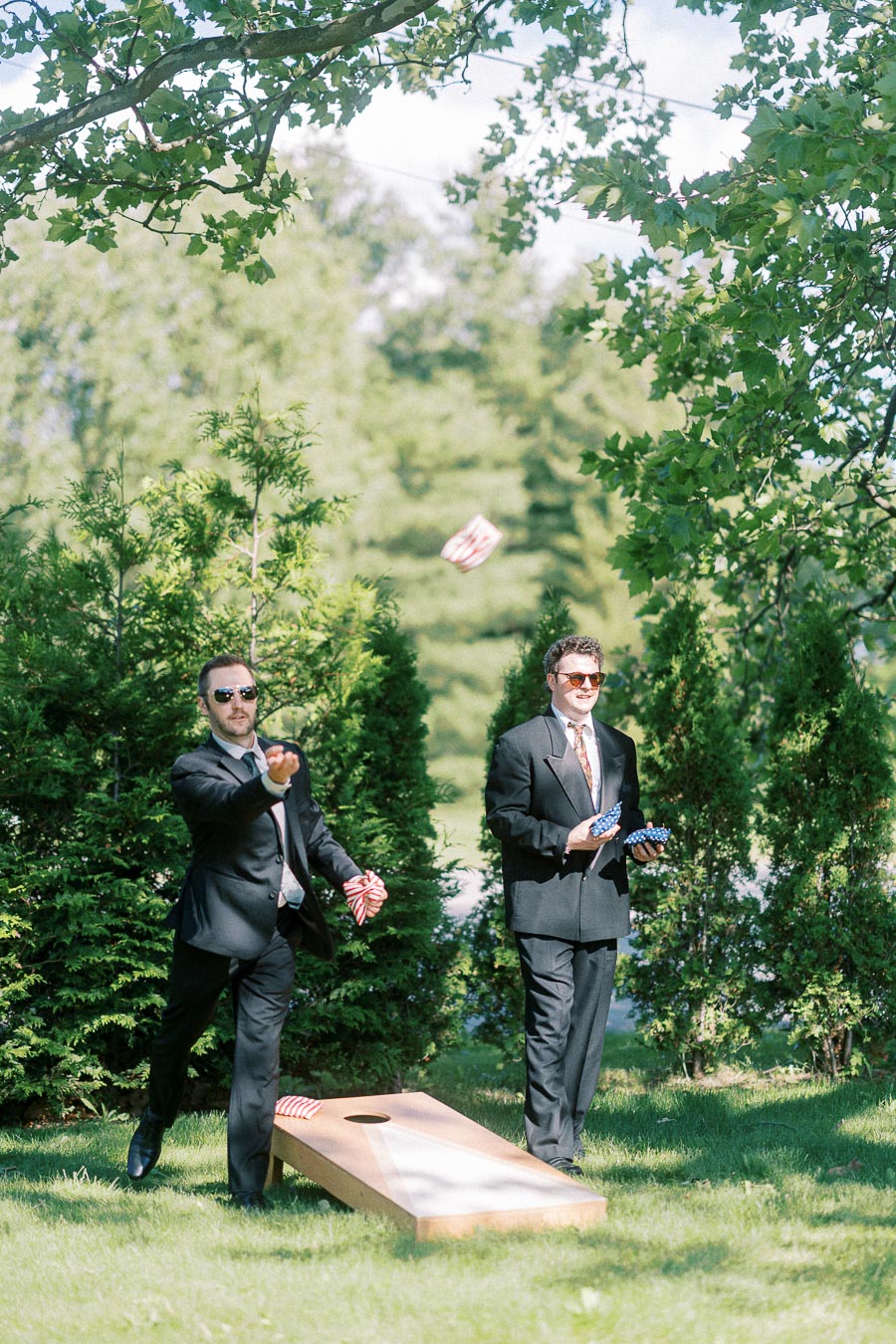 Two men in suits playing cornhole outdoors on a sunny day, surrounded by greenery, with one man tossing a red-striped bean bag towards the wooden board.