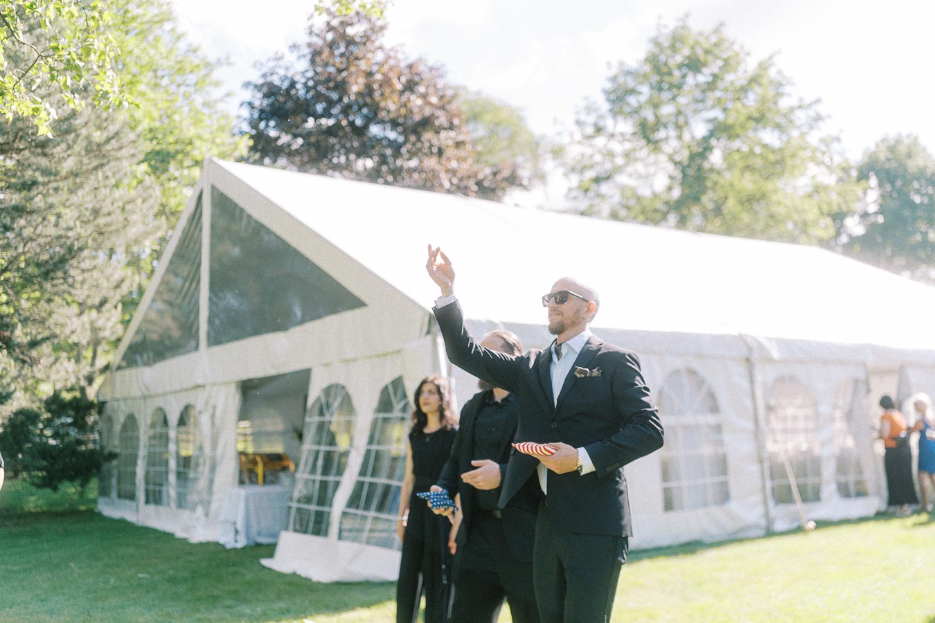 A man in a suit and sunglasses stands outdoors, engaged in a game, holding a colorful bean bag. He is in front of a large white event tent, surrounded by others in formal attire, amidst a sunny garden setting.