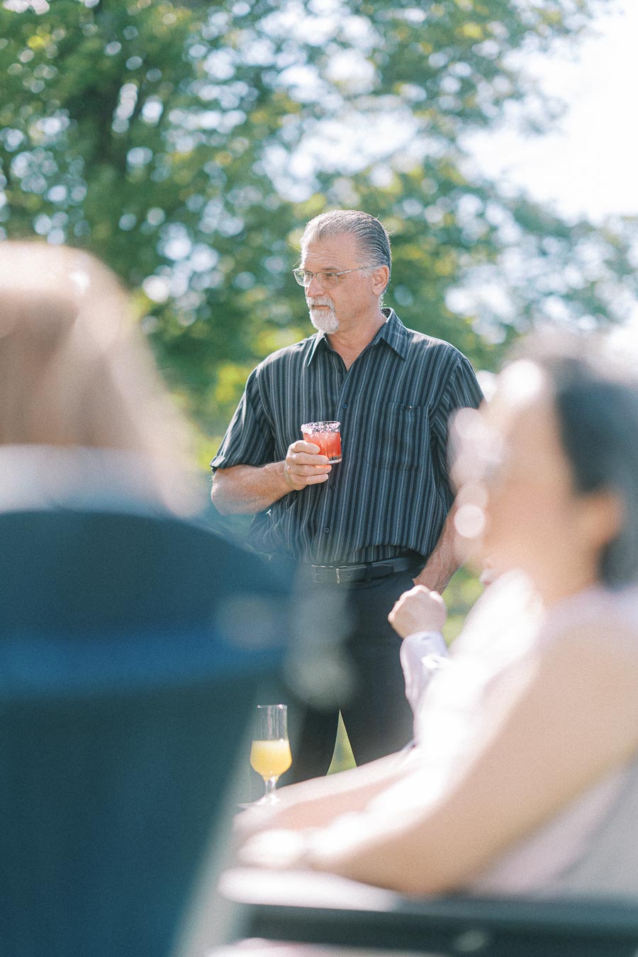 Man holding a drink while socializing outdoors at a sunny gathering, with blurred guests in the foreground and trees in the background.