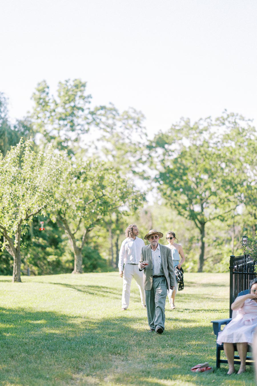 Senior man in a hat walking through a grassy park with two people following, while a person sits on a bench nearby.