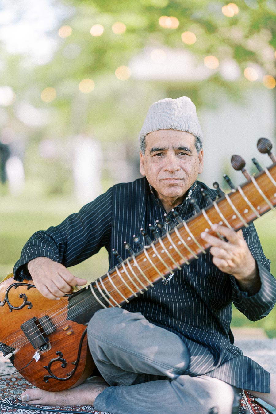 Elderly man playing a sitar outdoors, wearing a traditional cap and striped shirt, surrounded by blurred greenery in the background.