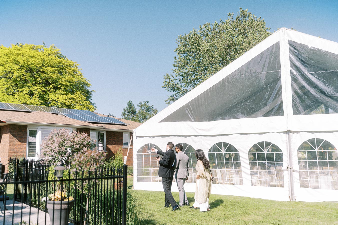 Three people walking beside a large white outdoor tent set up for an event, with solar panels visible on a nearby brick house roof, surrounded by lush greenery and clear blue sky.