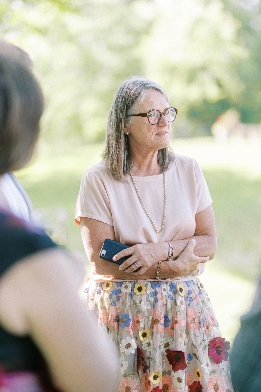 Senior woman with glasses holding a smartphone, wearing a floral skirt in a sunny outdoor setting.