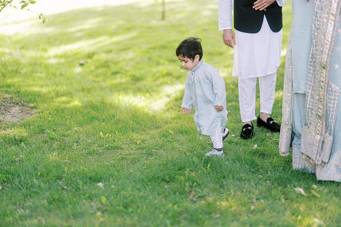 Toddler in traditional outfit walking on green grass, accompanied by adults in similar attire, with sunlight creating a serene outdoor setting.