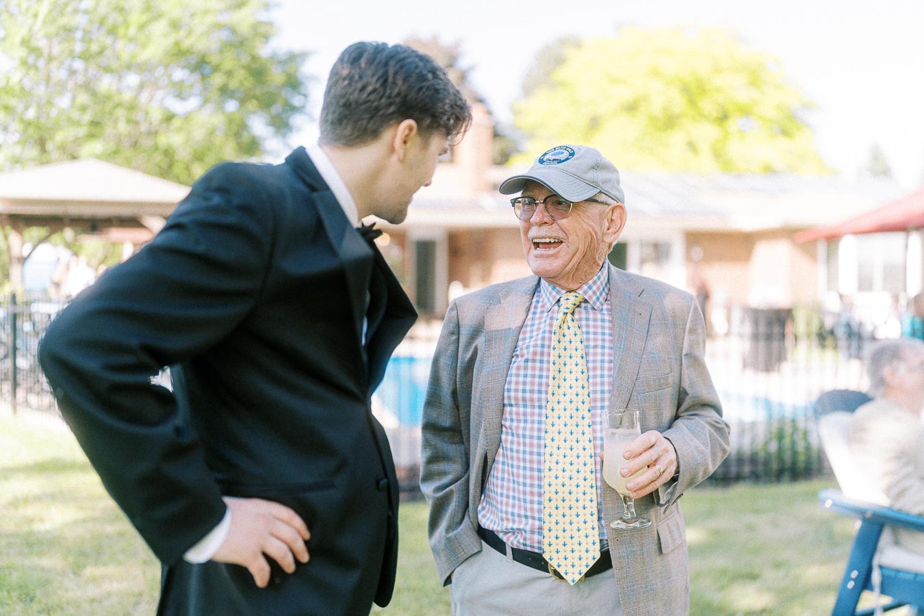 A young man in a black suit engages in conversation with an older gentleman in a checked shirt and tie, holding a drink, during an outdoor gathering on a sunny day.