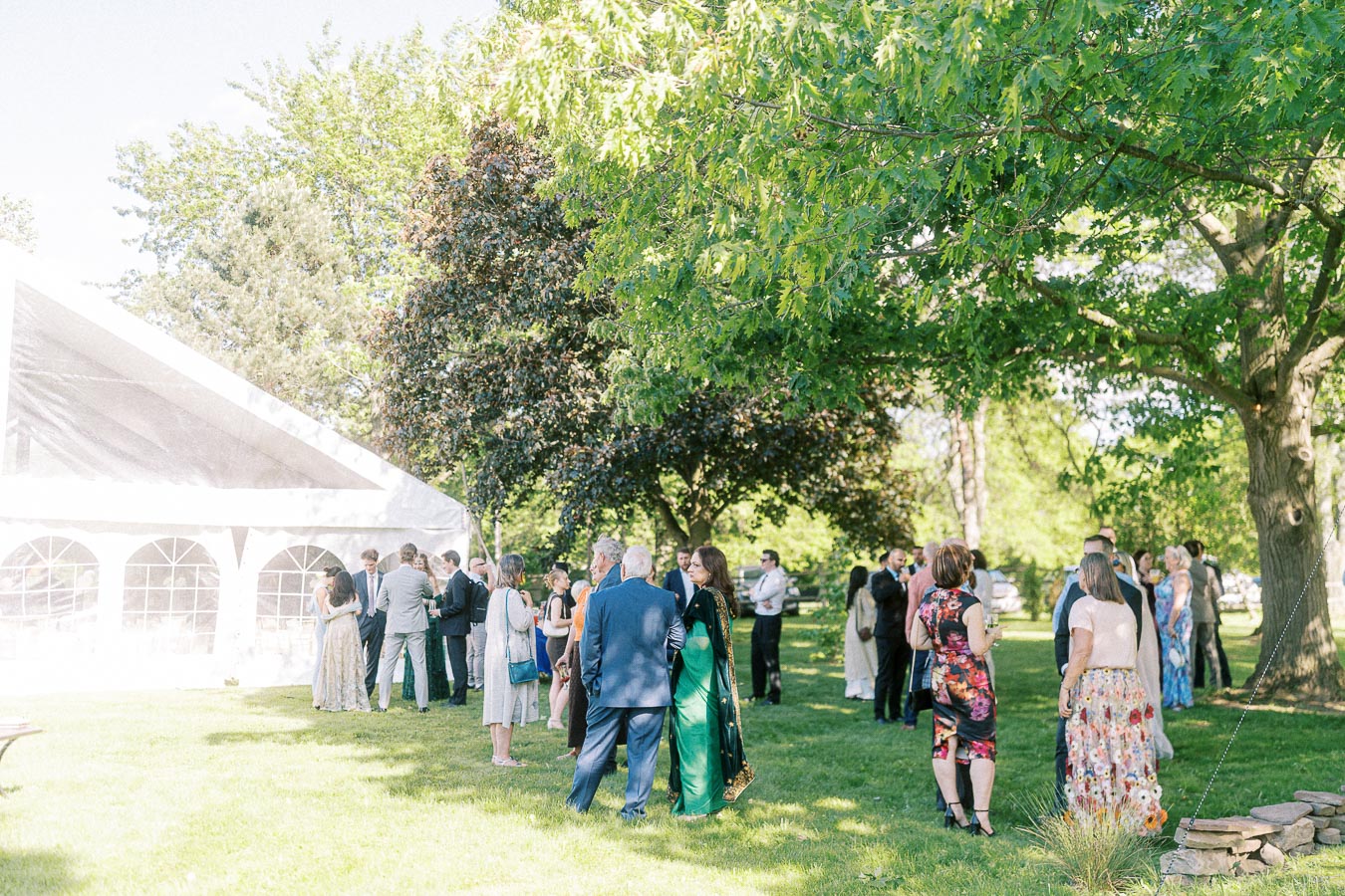 Outdoor wedding reception with guests socializing under trees beside a white event tent on a sunny day.