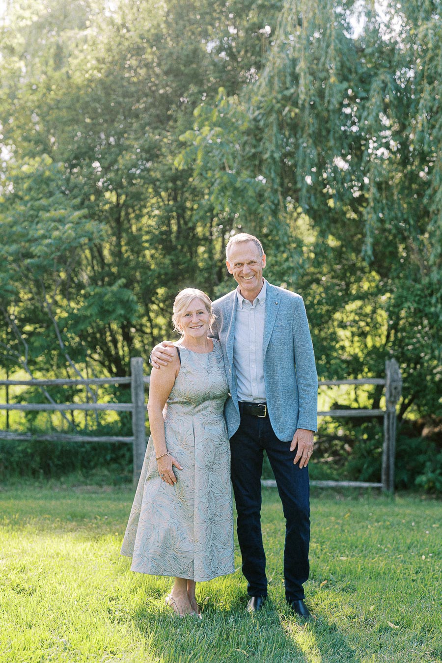 Elderly couple smiling in a sunny garden setting, with green trees and a wooden fence in the background.