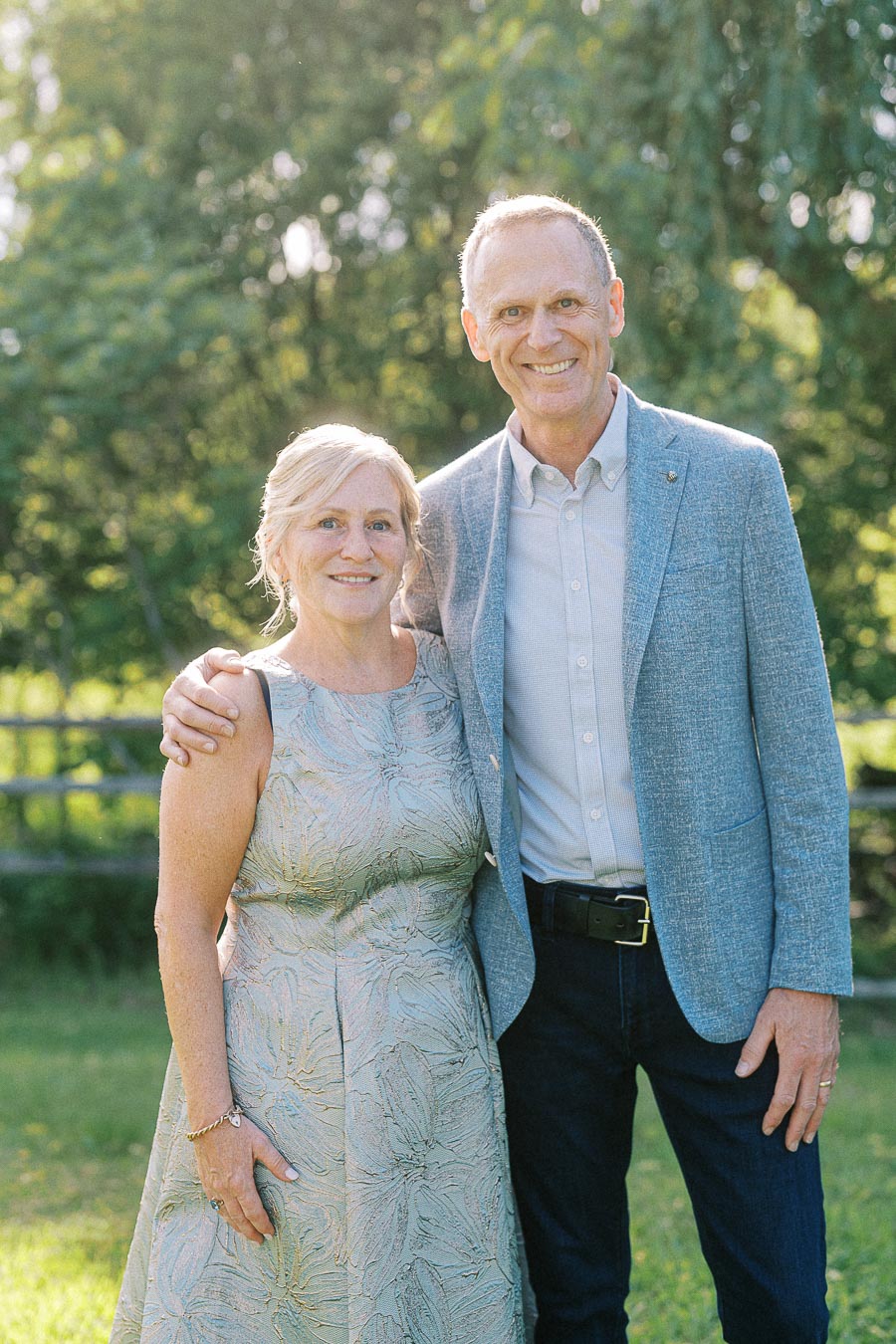 Elderly couple posing outdoors in elegant attire with lush green foliage in the background.