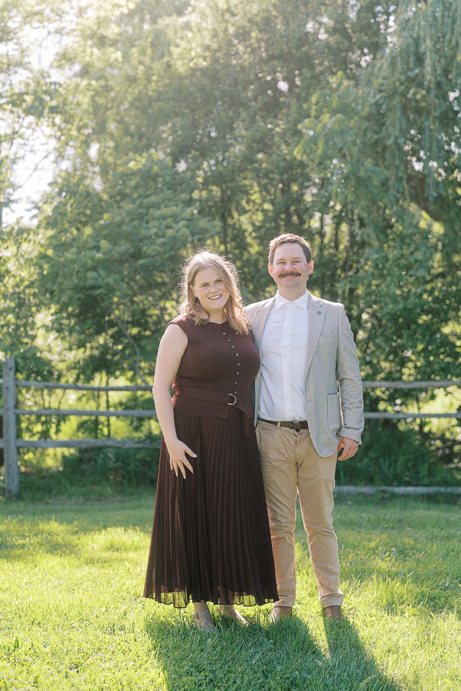 A smiling couple standing together in a sunny garden setting, featuring lush green trees and a wooden fence in the background. The woman is wearing a dark dress, and the man is dressed in a light blazer and khaki pants.