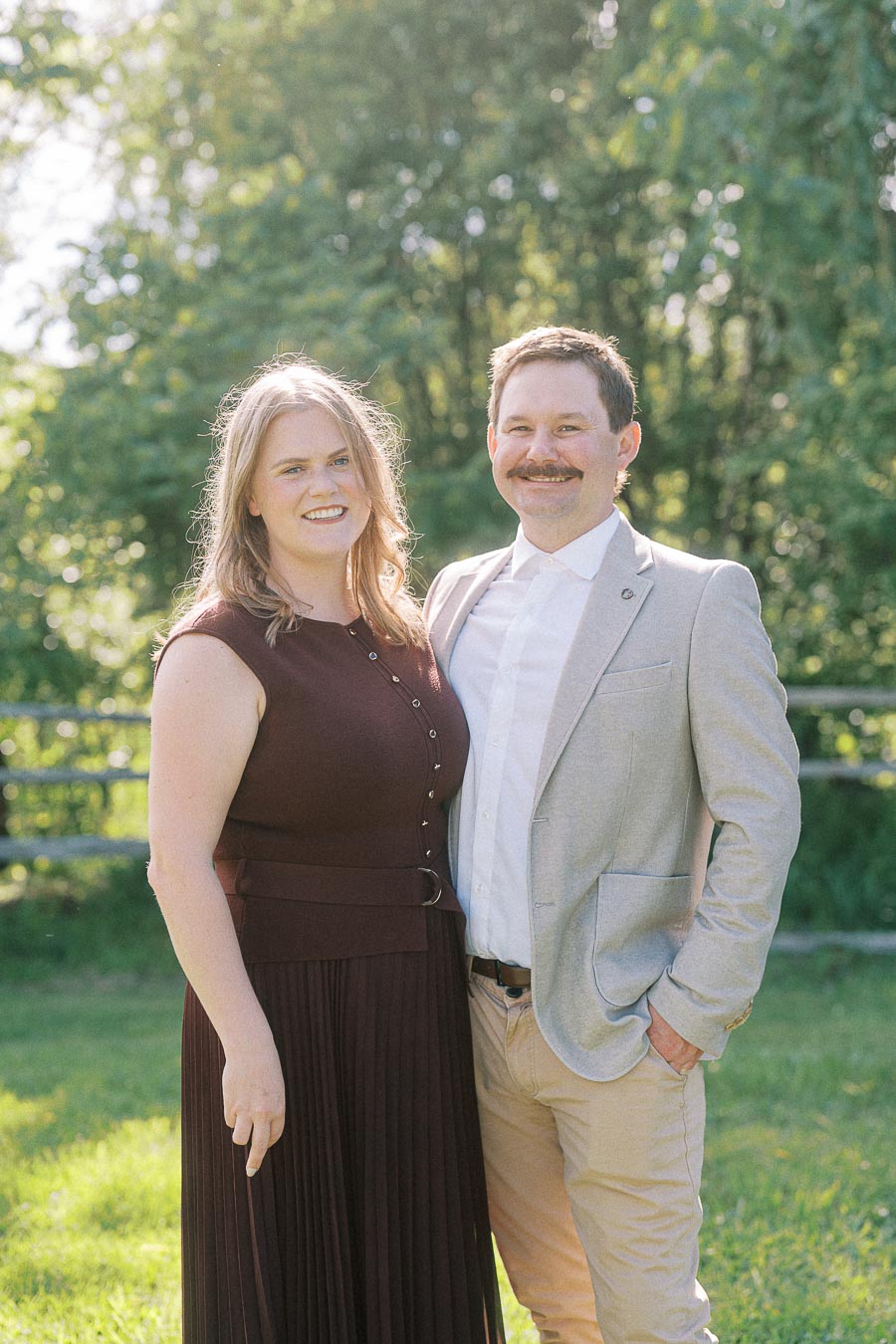 A smiling couple standing together in a sunlit garden, with lush green trees in the background. The woman is wearing a maroon dress and the man is dressed in a light gray blazer and beige pants.