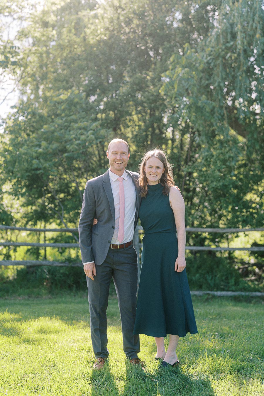 A couple posing outdoors in formal attire, with a lush green backdrop of trees and grass under sunlight. The man wears a gray suit with a pink tie, while the woman is in a sleeveless dark dress, both smiling warmly.
