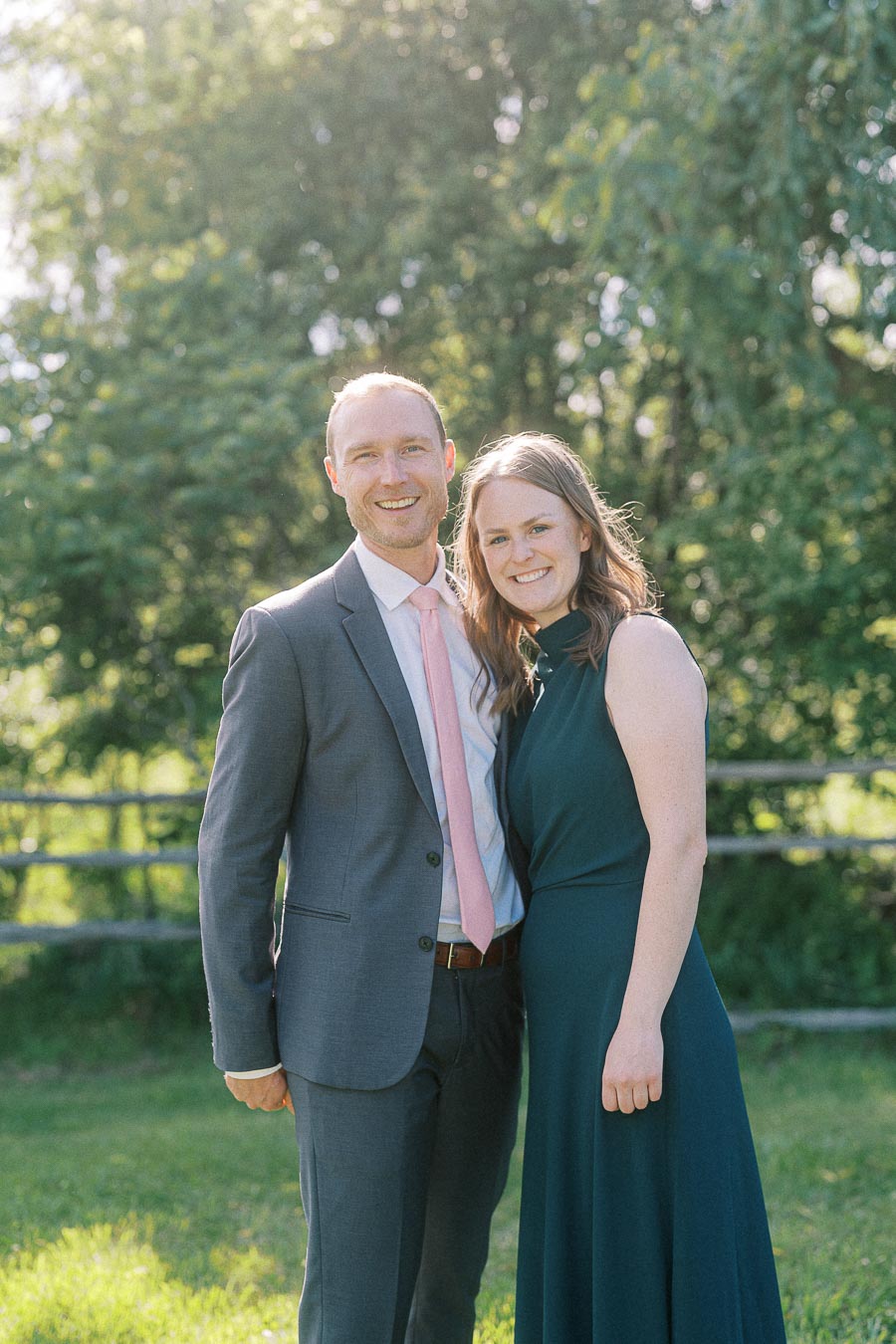 A smiling couple posing outdoors during a sunny day, with lush green trees in the background. The man is wearing a gray suit with a pink tie, while the woman is dressed in a dark green dress. They appear happy and relaxed in a natural setting.