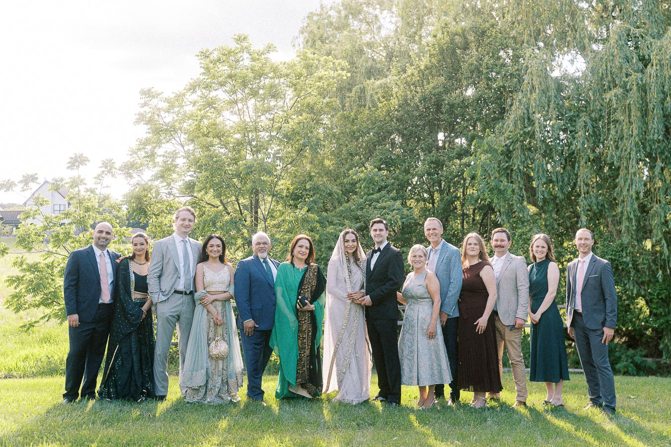 Group of elegantly dressed people posing outdoors at a garden wedding, with lush greenery and a bright sunny day as the backdrop.