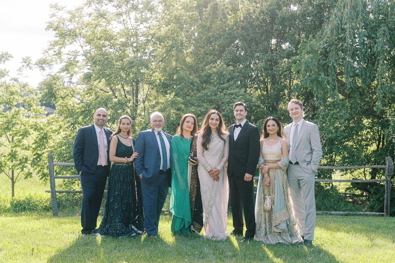 A group of elegantly dressed people standing outdoors in a lush green setting on a sunny day, showcasing formal and traditional attire, with trees in the background.